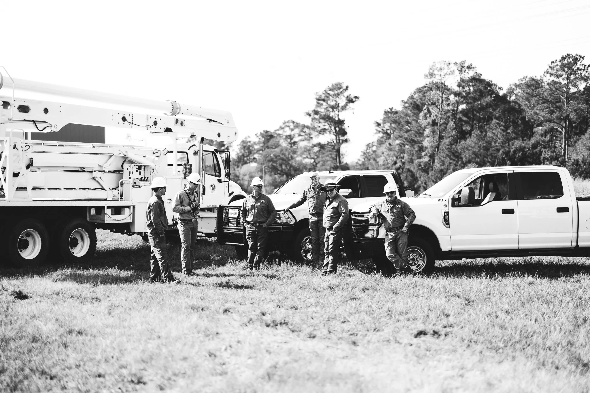 Group of workers wearing safety helmets standing near utility trucks in grassy field with trees in the background.