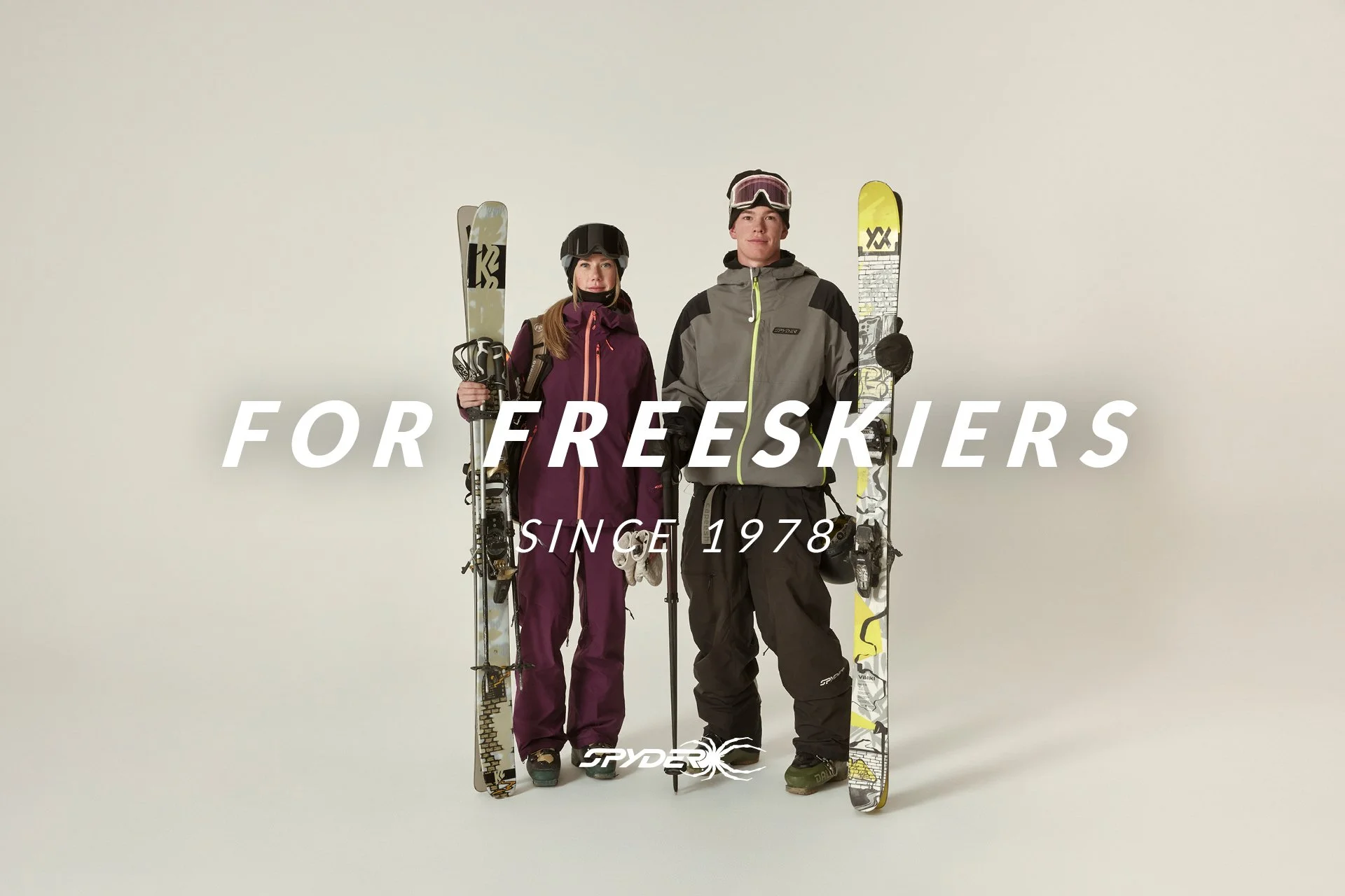 Two skiers, a woman in a purple jacket and a man in a gray jacket, holding skis and poles, standing against a plain background with the text 'For F reeskers Since 1978' and the Spyder logo.