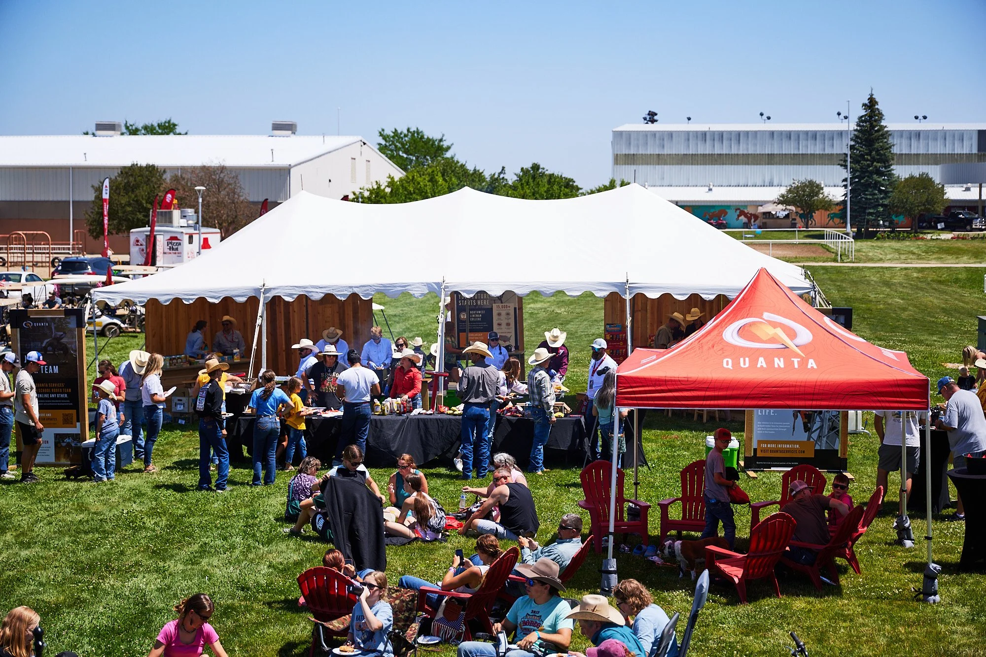 Outdoor community event on a grassy field with several tents, including a red Quanta tent, and a crowd of people, some seated and some standing, enjoying food and activities on a sunny day.