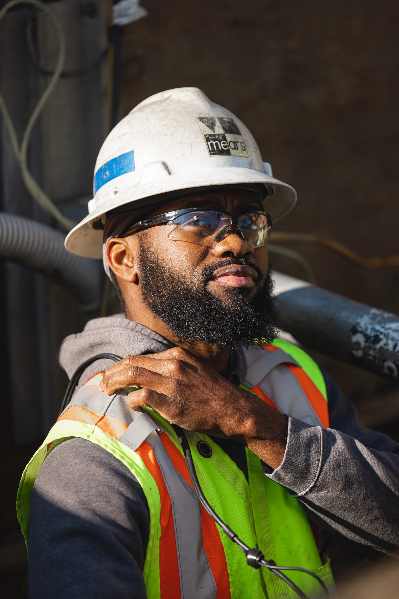 A construction worker wearing safety glasses, a white hard hat, and a neon safety vest, with a serious expression, holding a pipe or pole on his shoulder.