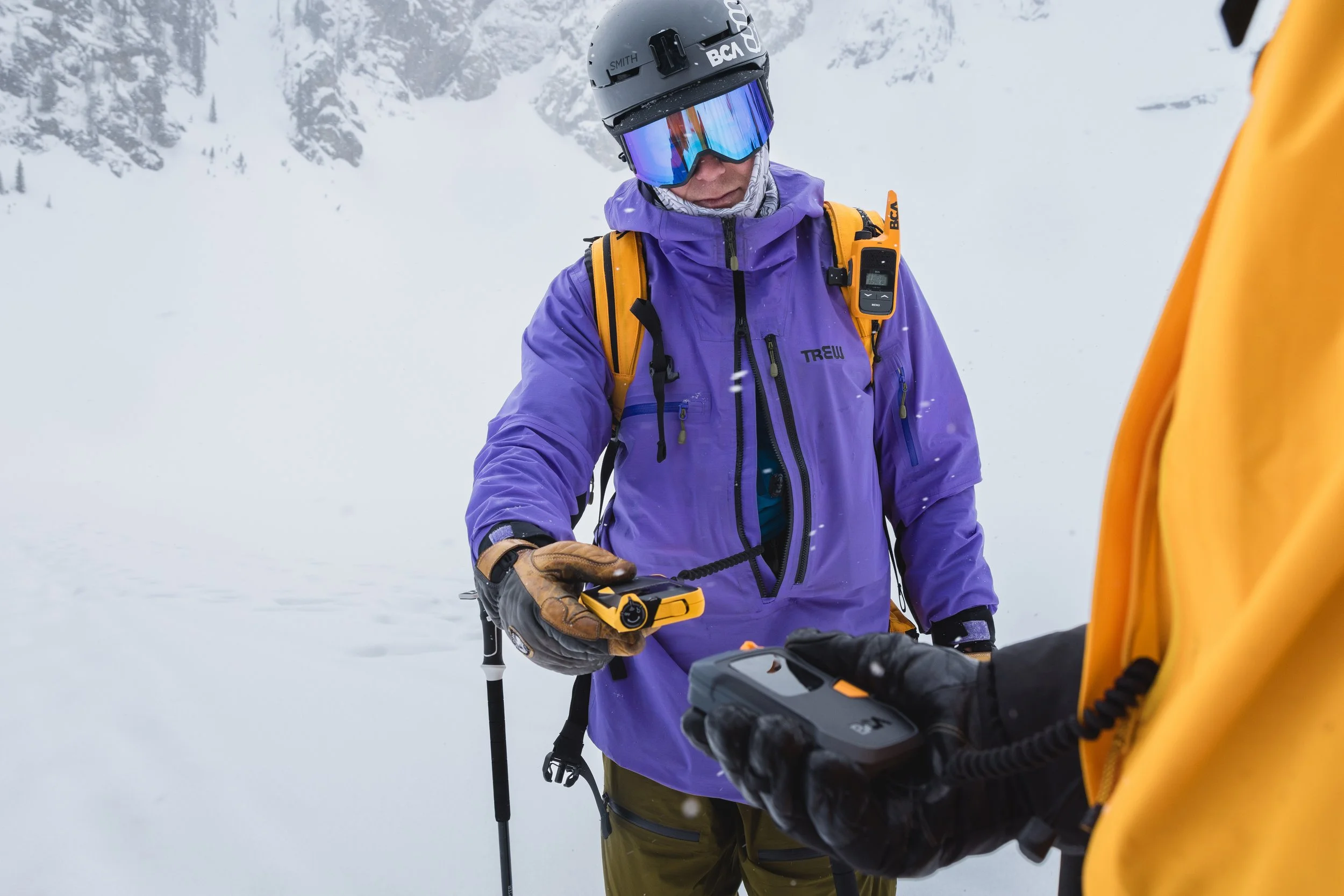 A person in winter gear, including a purple jacket, helmet, and goggles, holding a device connected to another person's handheld device in a snowy mountain landscape.