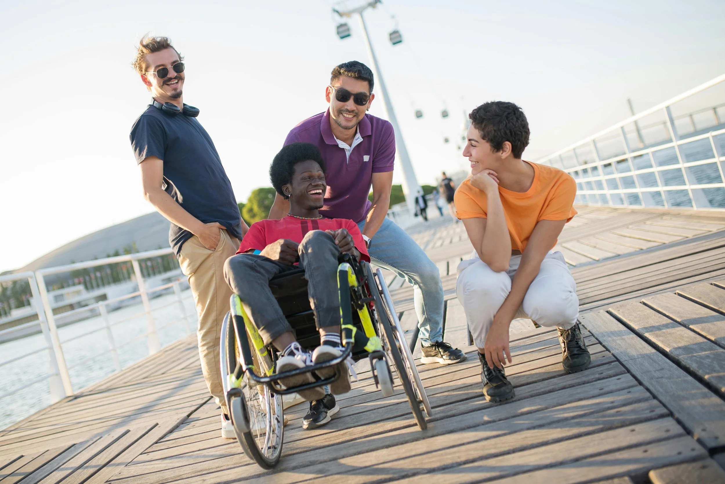 Four friends enjoying a sunny day on a wooden pier by the water, with one person in a wheelchair laughing and the others smiling and talking.