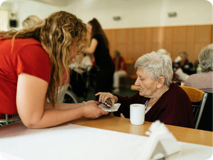 Young woman showing something on her phone to an elderly woman sitting at a table in a communal setting.