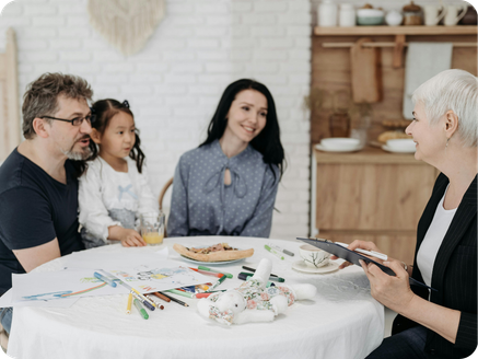 Family gathered around a table with art supplies, a pastry, and a toy, talking to an older woman holding a tablet.