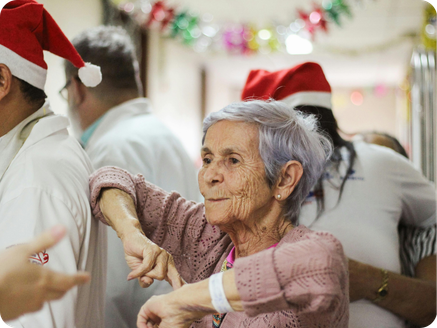 An elderly woman with gray hair wearing a pink sweater and a red Santa hat is dancing at a holiday celebration with other people in festive attire, including Santa hats.