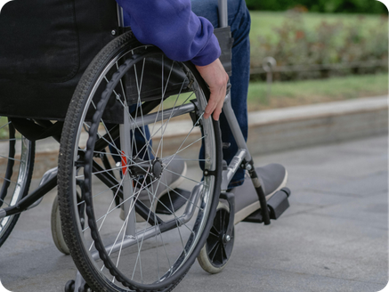 Person in a wheelchair on a sidewalk, with part of the wheelchair and a person’s hand visible.