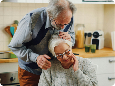 An elderly couple in a kitchen, with the man comforting the woman who is sitting at a counter, appearing distressed.
