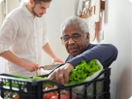Elderly man with gray hair and glasses reaching into a grocery cart filled with vegetables while a caregiver arranges items on a countertop in a bright kitchen.