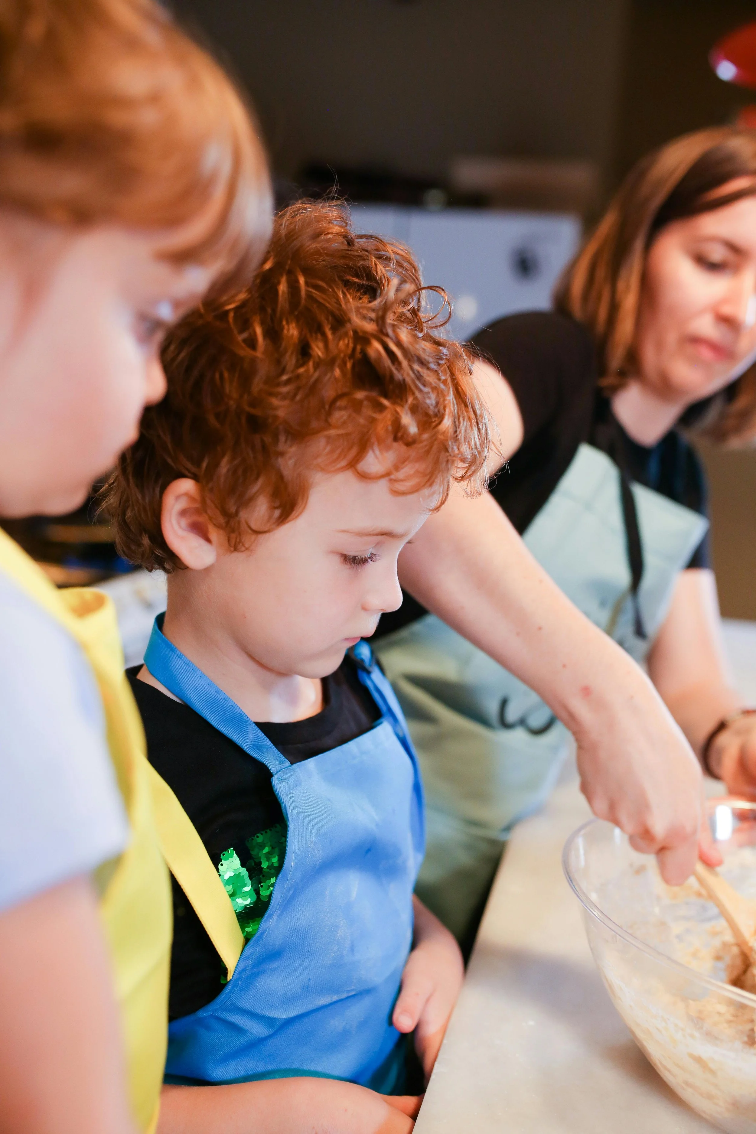 A woman and two children stand at a kitchen counter, baking or cooking together. The woman is serving or mixing ingredients in a large bowl, while the children watch attentively.