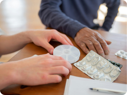 Person receiving a medicine reminder device from another person at a table with medication blister packs and a pen.