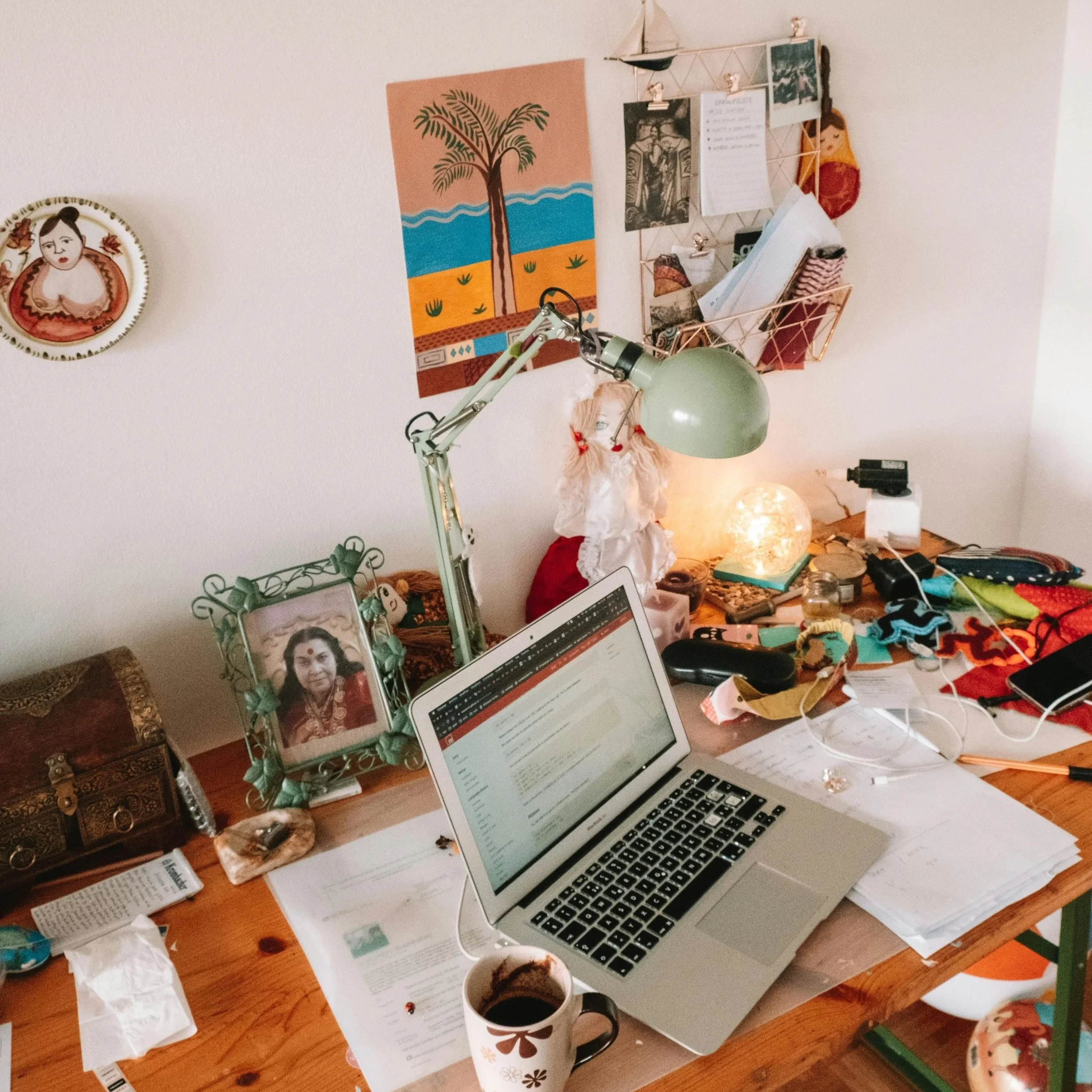 picture of a cluttered desk. theres a computer in the center of the desk. lots of trinkets around the desk and many small pictures attached to the wall behind the desk.
