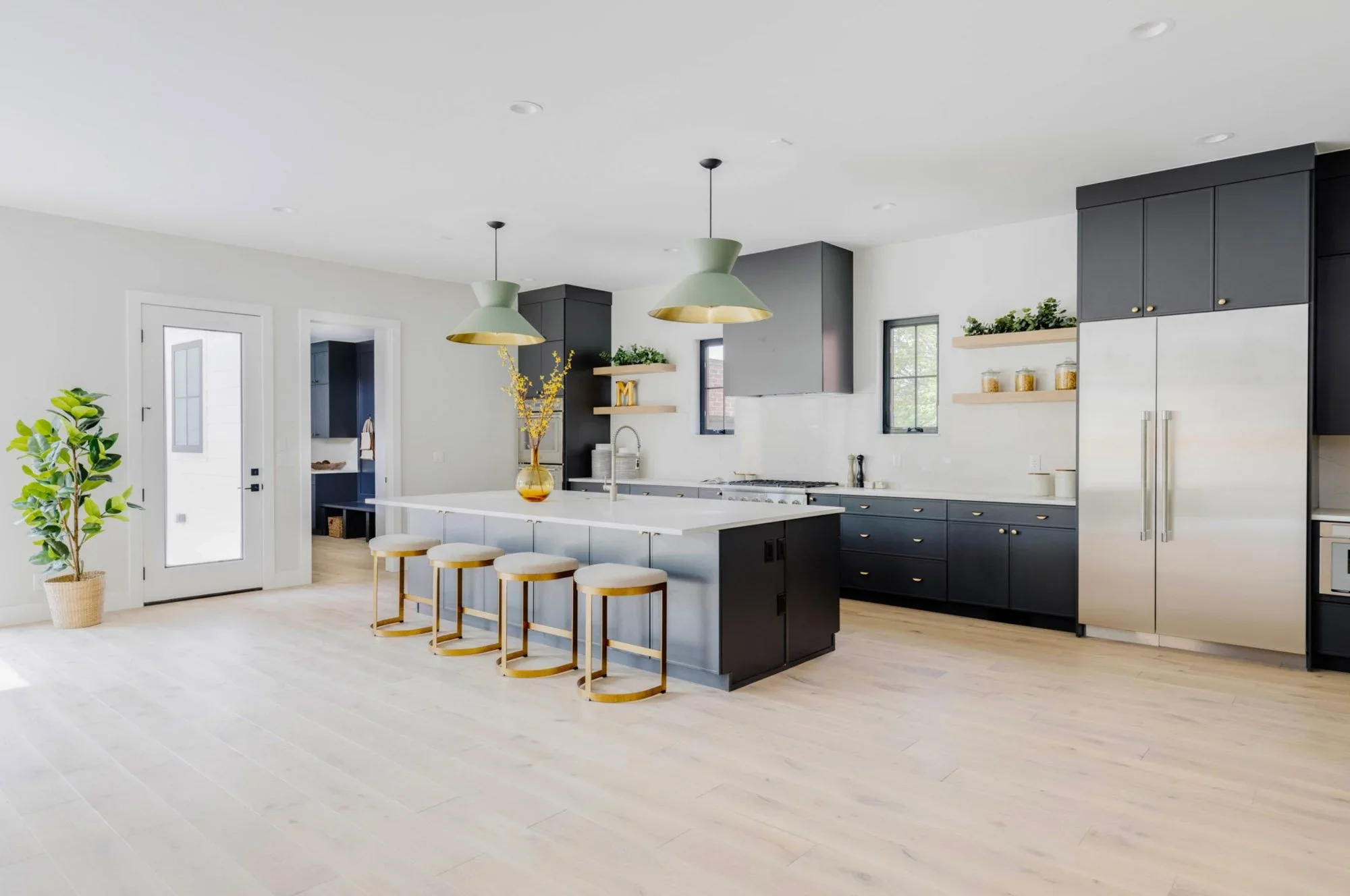 Modern kitchen with black cabinets, stainless steel refrigerator, white countertops, two green pendant lights, four bar stools, and minimal decor.