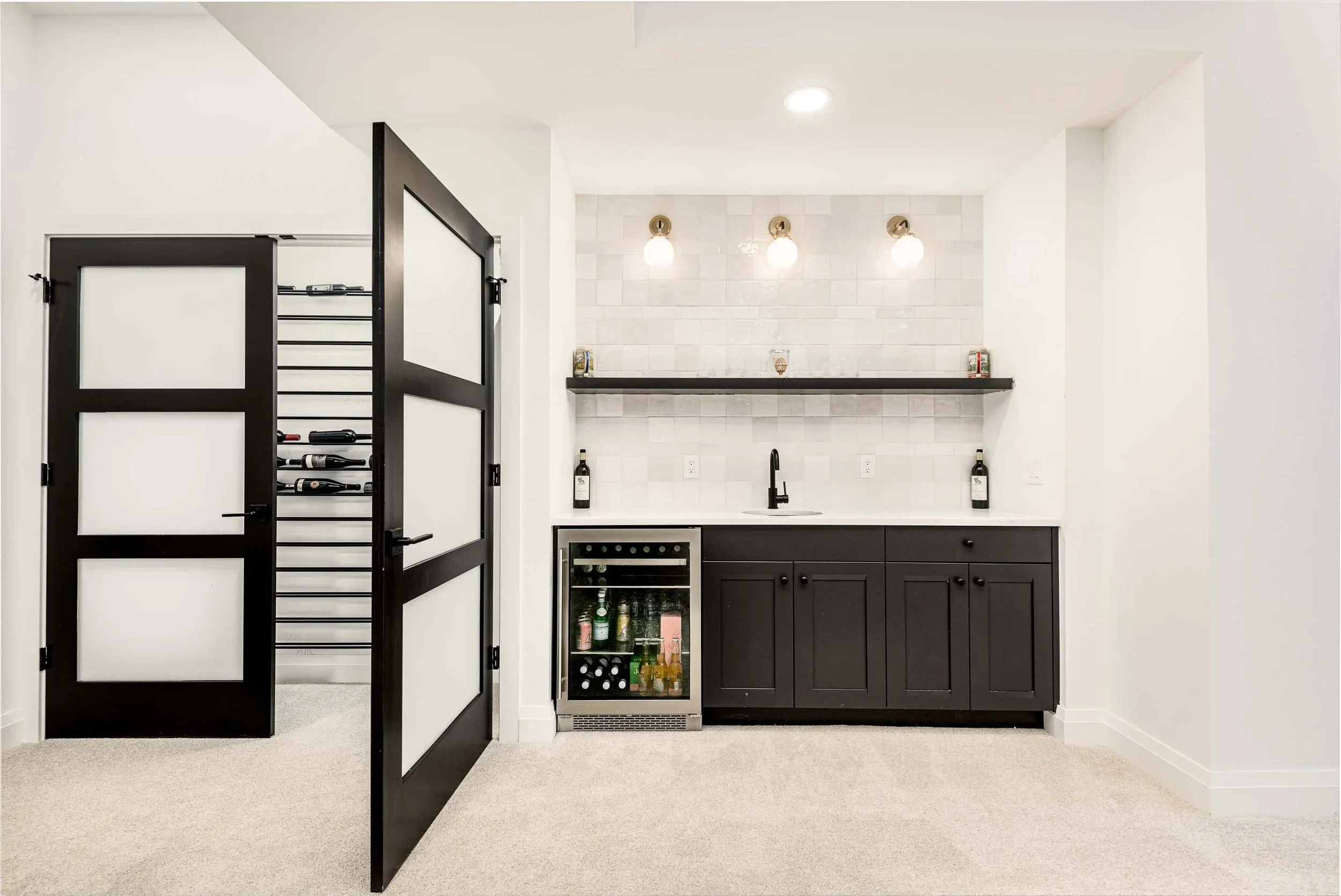 Mini bar area with black cabinets, white countertop, open shelf with small jars, wine bottles, a small wine cooler, and three ceiling light bulbs.
