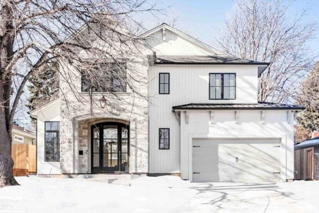 A modern, white two-story house with a black front door, large windows, and a two-car garage, surrounded by snow and leafless trees.