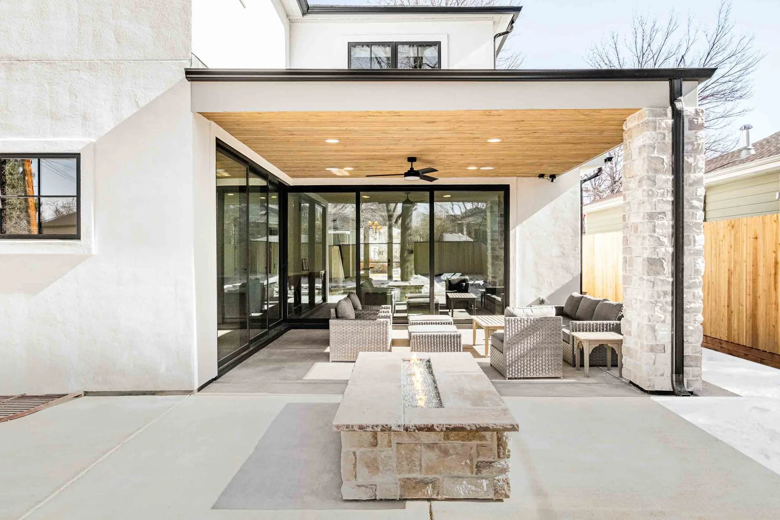 Modern outdoor patio with seating area, fireplace, and glass sliding doors leading into the house, under a wooden ceiling with ceiling fan.
