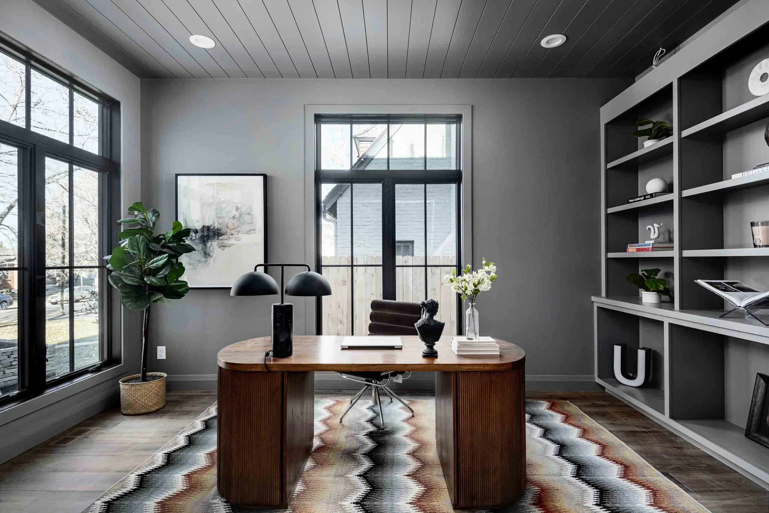 Modern home office with large windows, a wooden desk, a black office chair, a bookshelf with decorative items, a potted plant, and a colorful rug.