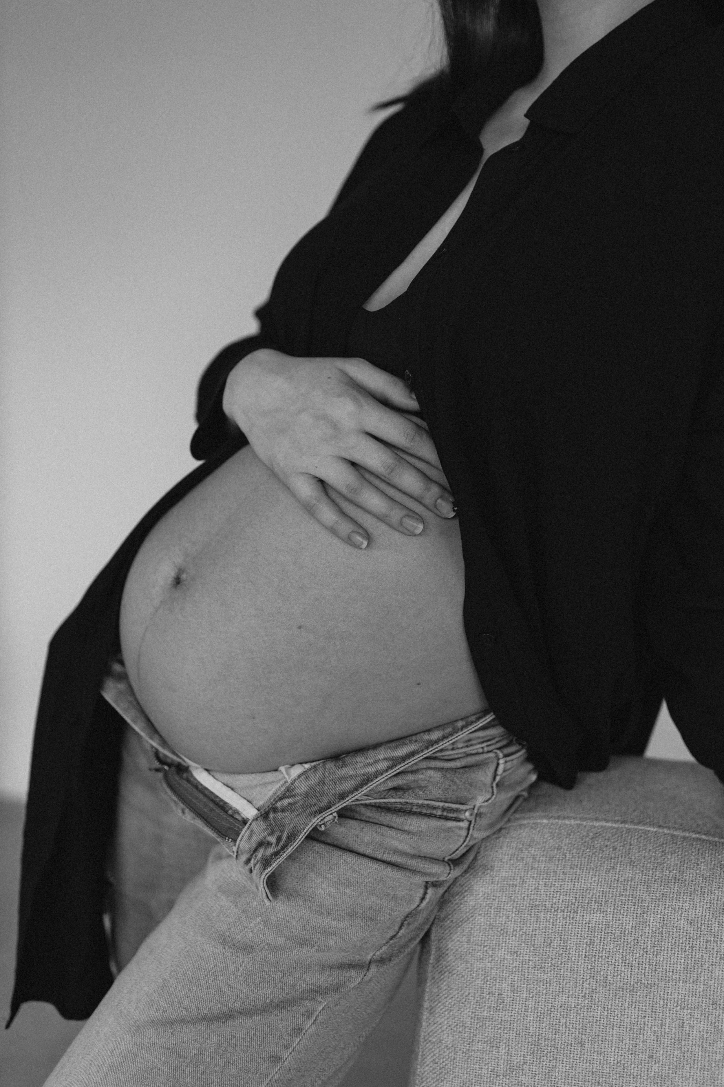 A black-and-white photo of a pregnant woman sitting on a sofa, holding her belly with one hand, wearing a black top and jeans.