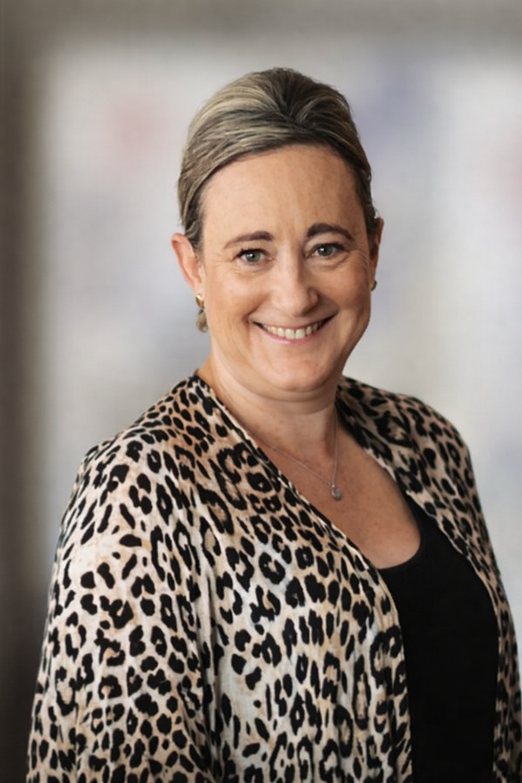 A woman with short blonde hair, wearing a leopard print jacket, black top, and a necklace, smiling at the camera.
