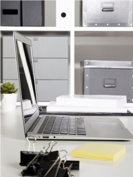 A modern workspace with a silver laptop, a small potted succulent, a stack of white papers, a yellow sticky note pad, and black binder clips on a white desk, with organized storage boxes and shelves in the background.