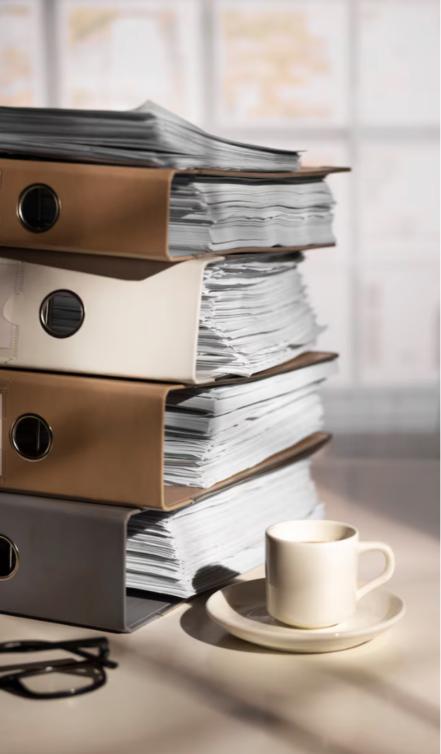 Stack of paper binders with loose papers on a desk, a white coffee mug on a saucer, and glasses, with a blurred window in the background.