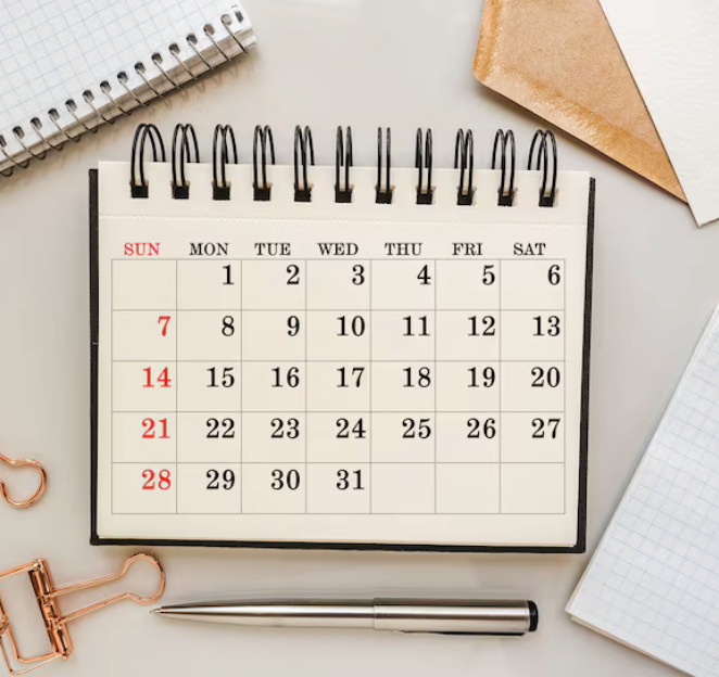 A desk calendar showing the month with dates and days, surrounded by notebooks, a pen, a paperclip, and a piece of brown paper.