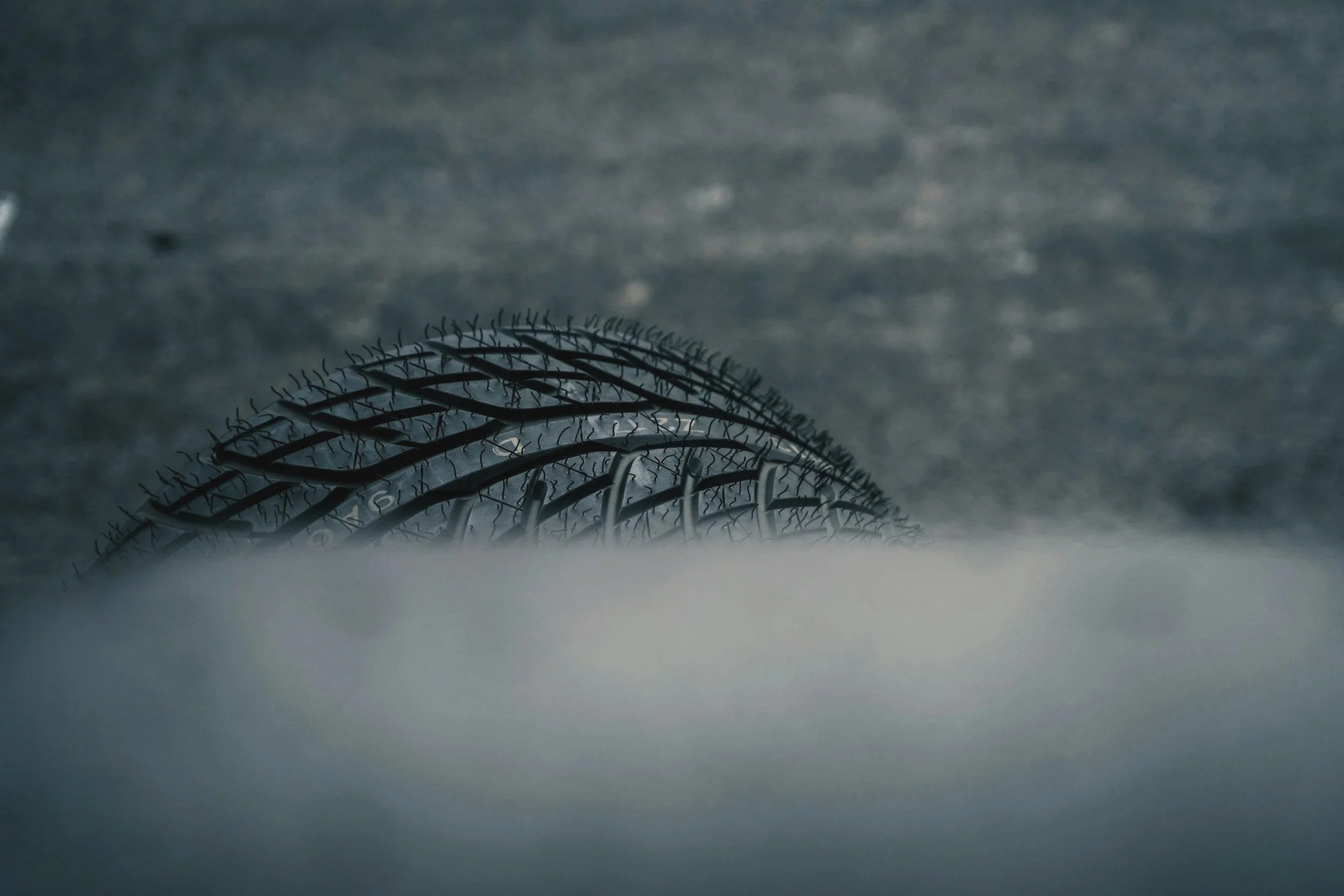 Close-up of a black rubber car tire partially covered by snow, with a textured gray asphalt road in the background.