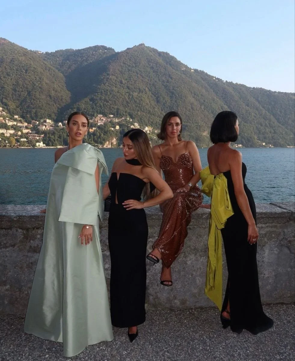 Four women in elegant dresses posing outdoors near a body of water with mountains in the background during sunset.
