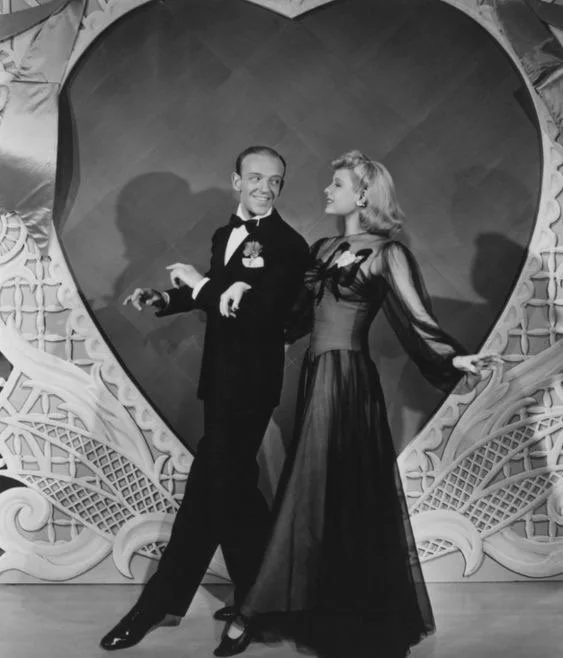 Man and woman in vintage formal attire dancing in front of a heart-shaped backdrop with ornate decorations.