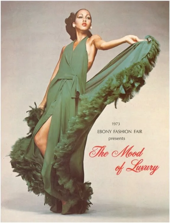 A woman in a green evening gown with a high slit, holding the dress's train. The dress has feathered trim. The text reads "1973 Ebony Fashion Fair presents The Mood of Luxury."