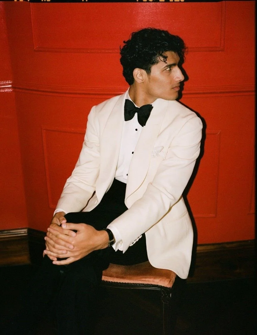 A man in a tuxedo with a black bow tie and white jacket sitting on a pink upholstered chair against a red wall, looking to the side.