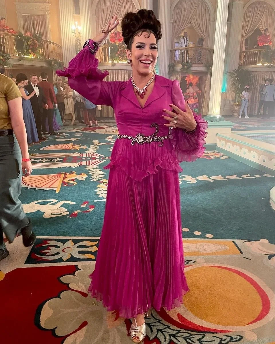 A woman in a vibrant pink vintage-style dress with ruffled sleeves and pleated skirt, smiling and waving at a formal event in a grand ballroom with ornate architecture and other guests in the background.