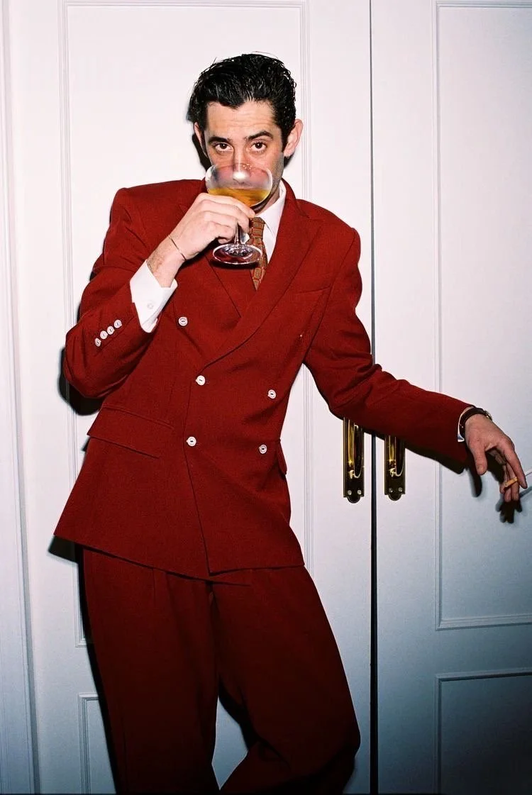 A man with dark, slicked-back hair is wearing a red double-breasted suit with a white shirt and patterned tie. He is standing in front of a white door, holding a large glass of wine or champagne up to his face, partially covering his mouth, and looki