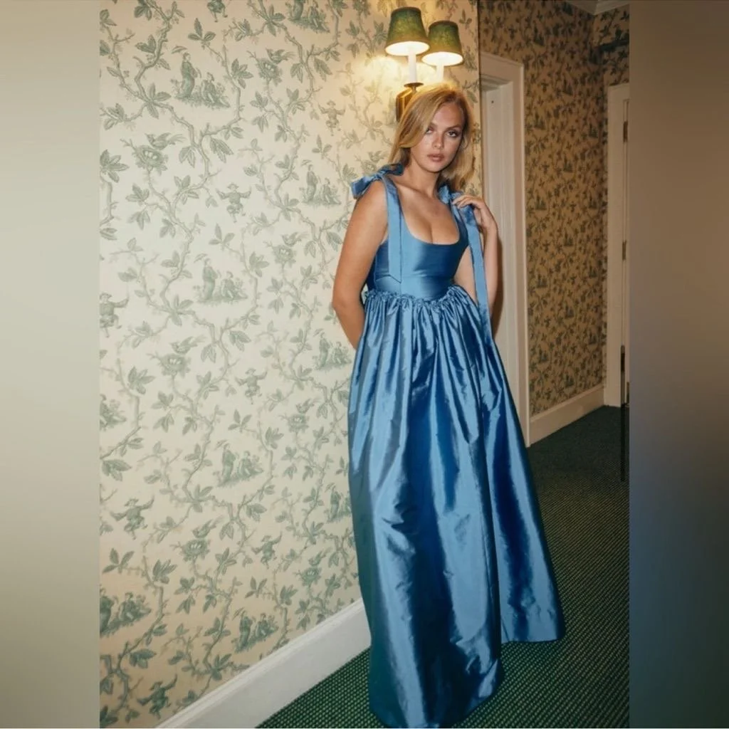 A young woman in a shiny blue gown with a full skirt and tied shoulder straps standing indoors against a floral wallpapered wall, illuminated by a wall sconce.