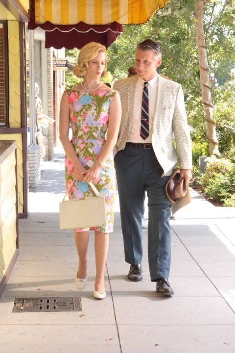 A woman and a man walking on a sidewalk outdoors, with trees and storefronts in the background. The woman is holding a beige handbag and wearing a colorful floral dress, while the man is dressed in a light-colored blazer, dark pants, a striped tie, a