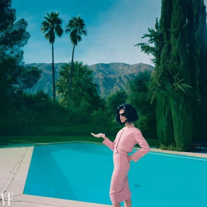 A woman in a pink dress and sunglasses stands by a pool, with a scenic backdrop of mountains and tall palm trees and greenery.