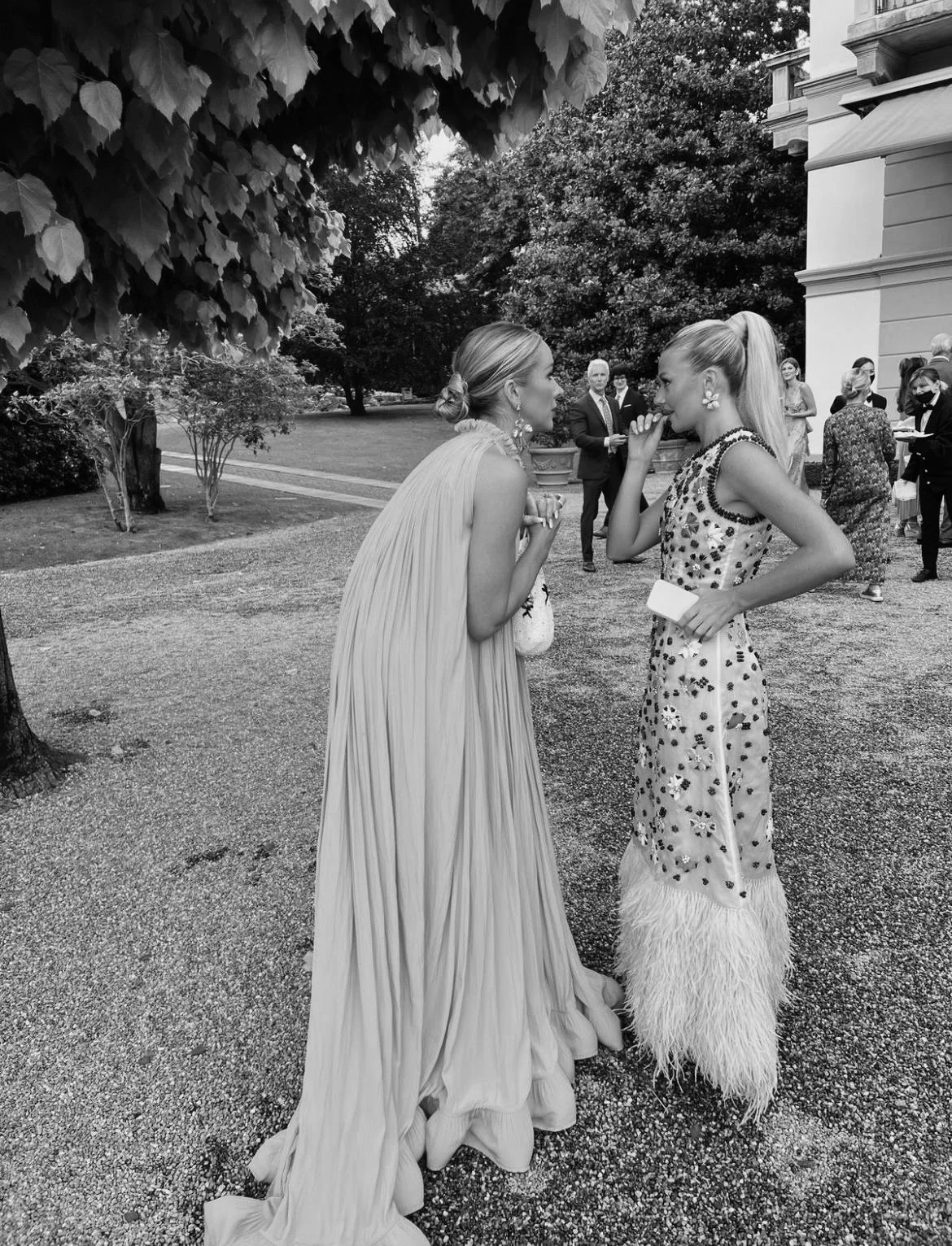 Two women are having a conversation outdoors at a formal event, with several other people in the background. One woman is wearing a long, pleated gown, and the other is wearing a patterned dress with feathered hem.