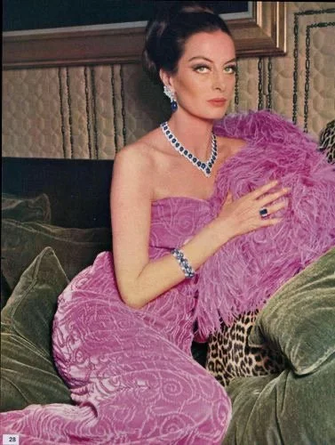 A woman with dark hair styled in an elegant updo, wearing a strapless purple gown and matching jewelry, sitting on a sofa with green cushions, holding a purple feathered fan, in a vintage-style room.