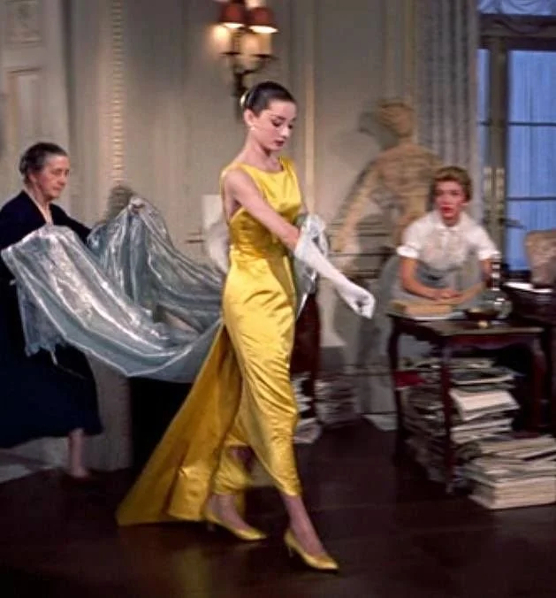 A woman in a yellow satin evening gown walking in a room with other women, one holding a silver fabric, indoors with classical decor and a window.