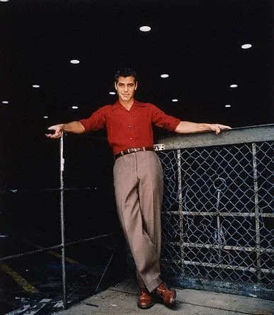 A man in a red shirt and beige pants leaning against a chain-link fence at night, with lights overhead.