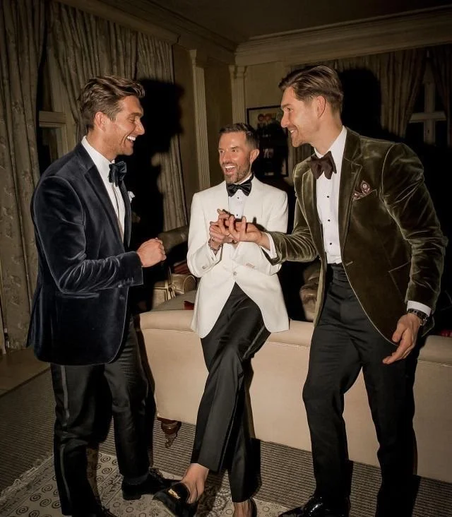 Three men in tuxedos dancing and laughing in a warmly lit room, with one man in a white tuxedo and the others in black and green velvet tuxedos.
