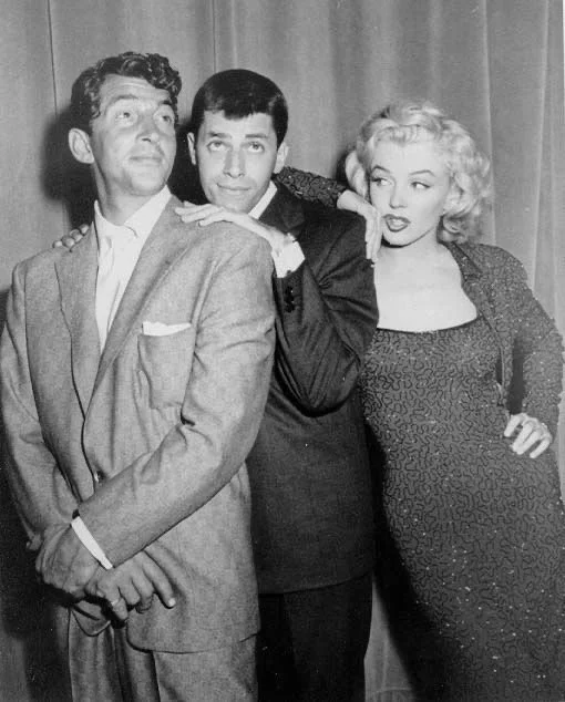 Black and white photo of three people posing together, with two men and one woman, in front of a curtain backdrop.