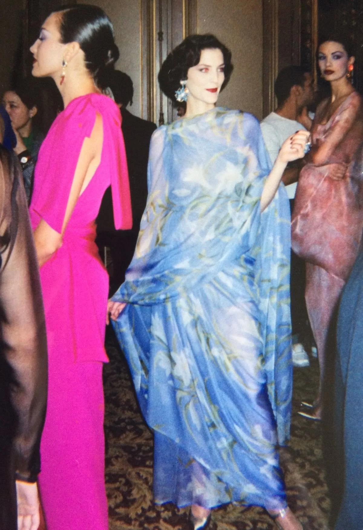 Women in fashionable dresses attending a social event, with one woman in a blue, patterned gown, and another in a bright pink dress, in an elegant room.