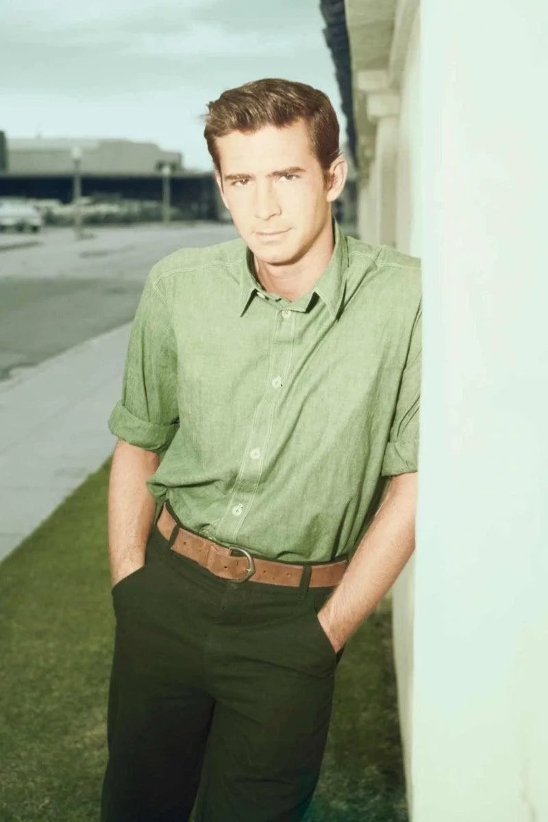 A young man with brown hair wearing a green button-up shirt and black pants, standing outdoors next to a white wall with his hands in his pockets.