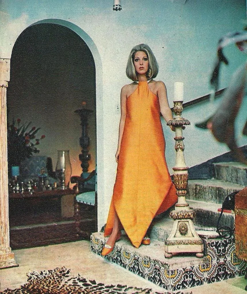 A woman with gray hair standing on a staircase next to a tall candle holder, wearing an orange halter dress.