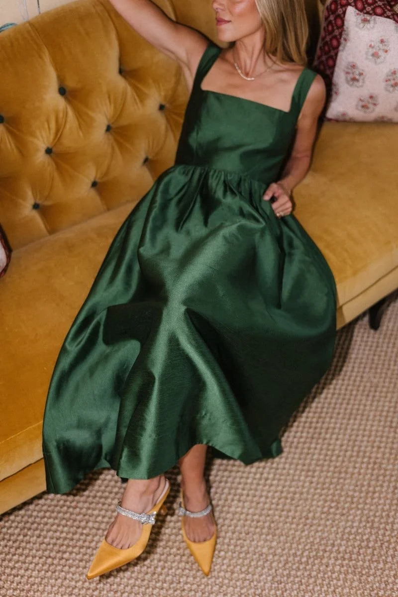 A woman in a long, shiny green dress sitting on a mustard-colored tufted sofa. She is wearing yellow shoes with embellishments and a pearl necklace, with her arm resting on the sofa back.