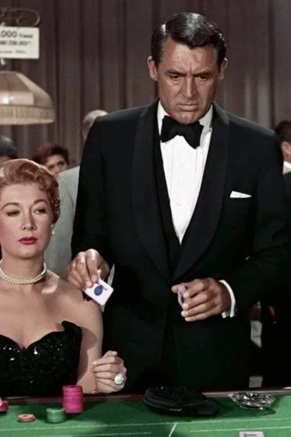 Scene from a casino, a woman with red hair and a pearl necklace is sitting at a gambling table with poker chips, next to a man in a tuxedo. The man is standing, holding some chips, and the woman is looking slightly downward, with a serious expression