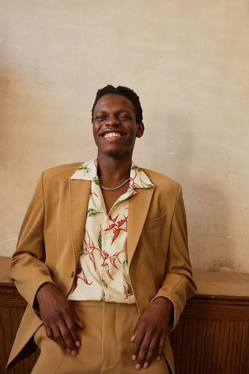 Young man smiling, wearing a tan blazer over a tropical shirt, standing against a plain wall.