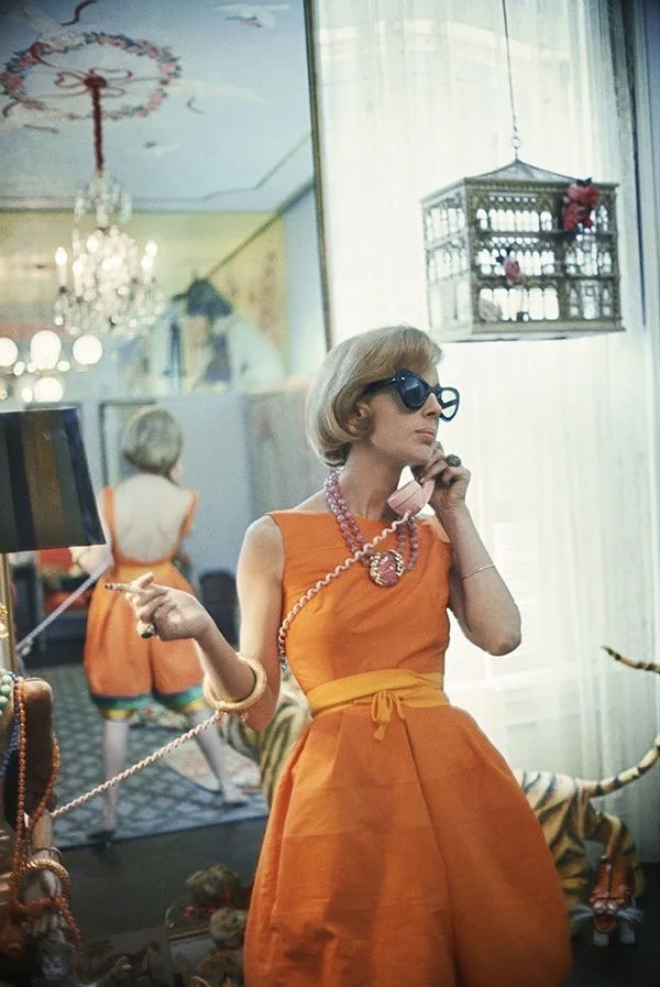 A woman in vintage orange dress with necklace and sunglasses talking on pink rotary phone inside a decorated room, with a child in similar orange outfit in the background and a birdcage hanging from the ceiling.