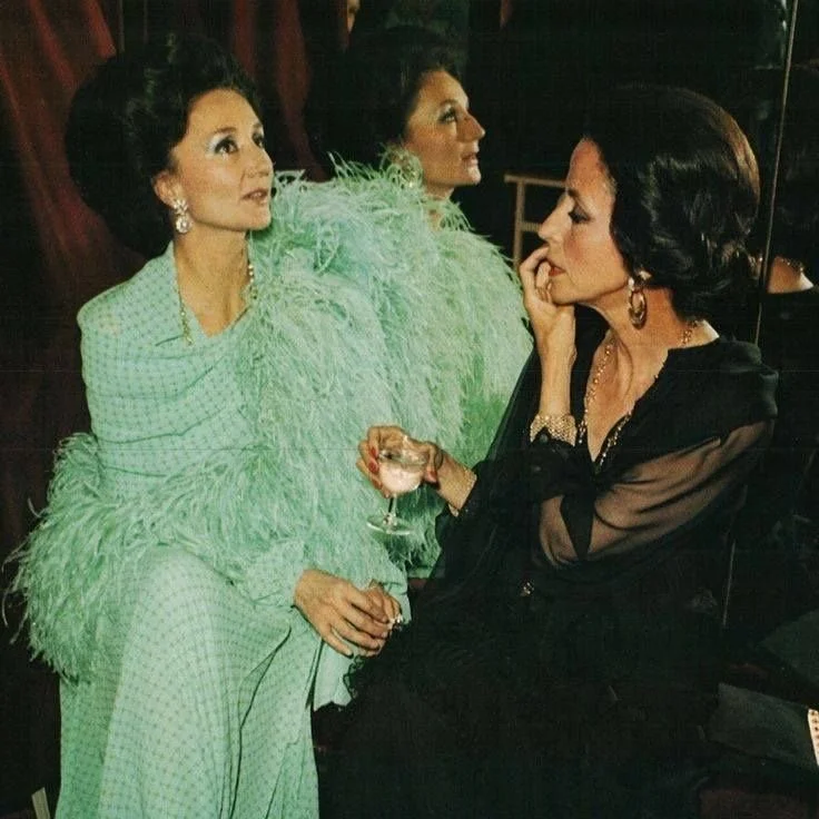 Two women dressed in elegant evening wear, one in a light green feathered gown holding a glass, conversing with a woman in black attire in a dimly lit room with dark wood furnishings and a mirror.