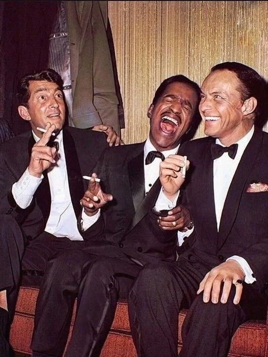 Three men dressed in tuxedos sitting together, enjoying a moment of laughter and conversation at a social event.
